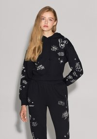 Felpa nera con cappuccio e pantaloni abbinati realizzati in un tessuto morbido, caratterizzati da stampe grafiche bianche di vari design, inclusi stelle e motivi di testo.