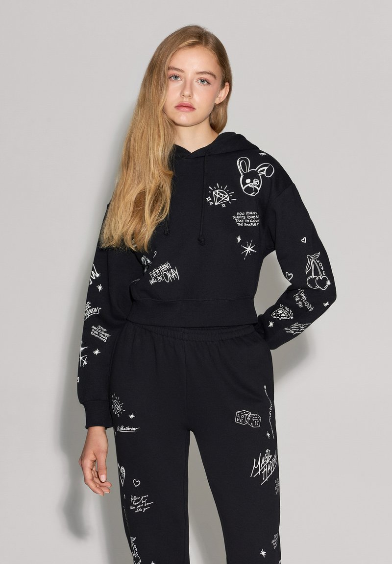 Felpa nera con cappuccio e pantaloni abbinati realizzati in un tessuto morbido, caratterizzati da stampe grafiche bianche di vari design, inclusi stelle e motivi di testo.
