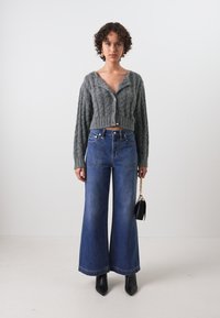 Cardigan en tricot gris, coupé, avec motif en cable ; jeans bleu taille haute, à jambe large ; sac à main noir ; bottines en cuir lisse.