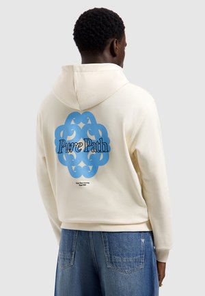 Man draagt een crèmekleurige hoodie met een blauw cirkelvormig ontwerp en de tekst "Pure Path" op de achterkant, gecombineerd met losse blauwe spijkerbroek.