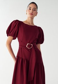 Bordeaux-kleurige jurk met pofmouwen, vierkante neklijn en een ceintuur met een gouden gesp. De rok heeft gelaagde ruches voor extra textuur.