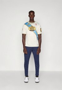 Hvit sportsdrakt med blå diagonal stripe, svart logo og korte ermer, kombinert med marineblå treningsbukser og hvite joggesko.