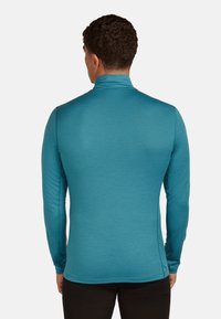 Man facing away wearing a fitted turquoise long-sleeve turtleneck shirt and black pants against a plain background.