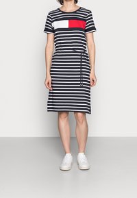 Gestreiftes Kleid in Marineblau und Weiß, mit einem roten Block auf der Brust, kurzen Ärmeln, einem Kordelzug an der Taille und einem knielangen Saum.