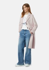 Manteau en laine rose clair avec deux boutons, associé à un t-shirt graphique blanc et un jean en denim à jambes larges. Les chaussures sont des baskets claires.