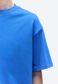 Személy egyszínű élénk kék rövid ujjú pólót visel egy világos háttér előtt, látható a felső mellkas és a jobb váll.