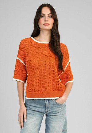 Femme aux longs cheveux foncés portant un pull en maille orange texturée avec un bord blanc et un jean bleu clair, debout avec une main dans la poche.