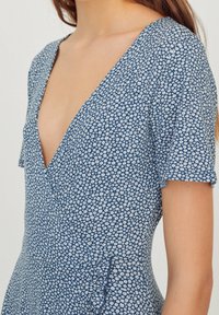 Modré květinové šaty s hlubokým véčkovým výstřihem, krátkými rukávy a strukturovanou tkaninou s malým vzorem bílých květin. Doplňují je volánky.