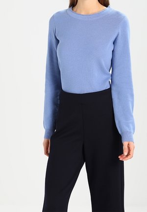 Woman wearing a light blue long-sleeve sweater tucked into high-waisted black pants, standing against a plain white background.