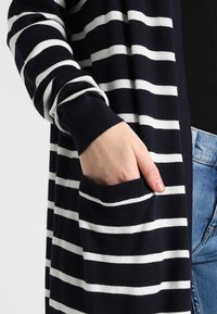 Mano en el bolsillo de un cárdigan a rayas en azul marino y blanco, llevado sobre una camiseta negra y unos vaqueros azules.