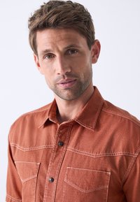 Rust-colored, textured shirt with a button-up front, two chest pockets, and contrasting white stitching. Features a classic collar and relaxed fit.