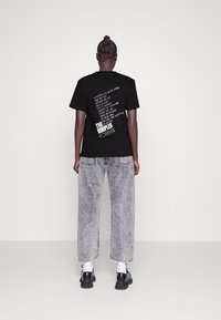 Camiseta de algodón negra con gráficos de texto blanco en la parte posterior. Combinada con unos jeans de mezclilla gris claro y holgados, y zapatos negros.