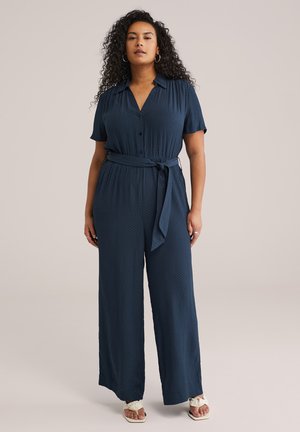 Moteris su garbanotais plaukais, vilkinti tamsiai mėlynos spalvos kombinezoną su diržu ir baltomis sandalais, stovinti priešais šviesų, vienspalvį foną.