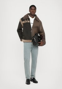 Braun- und schwarz gestreifter Wolljacke mit Schafsfellkragen, Reißverschluss und dekorativen Beschlägen, kombiniert mit einer hellen Jeans und einer schwarzen Handtasche.