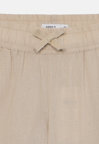 Pantalon en lin beige avec une taille élastique et un accent en nœud. Présente un tissage texturé et une coupe lâche pour le confort. Étiquette visible en haut.