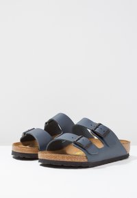 Grå syntetiske sandaler med to justerbare spenner, korkfotseng og teksturert svart såle. Enkel design egnet for hverdagsbruk.