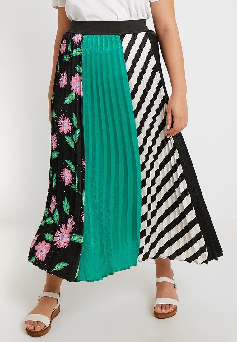 Femme portant une jupe midi plissée avec des panneaux fleuris noirs, turquoise et rayés diagonaux noir et blanc, associée à des sandales blanches.