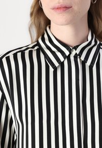 Svartvit randig blus med knäppkrage, slät tygstruktur och klassisk passform med vertikala linjer.