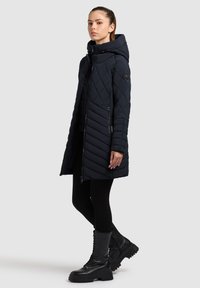 Navy gewatteerde lange jas met een capuchon, voorzien van een frontale rits, zijrits zakken en opvallende ruitenpatronen. Gecombineerd met zwarte leggings en laarzen.