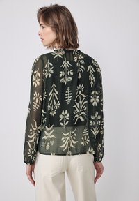 Transparent schwarze Bluse mit floralem Muster, langen Ärmeln, gerafftem Ausschnitt und leichtem Material. Sie weist cremefarbene botanische Designs auf.