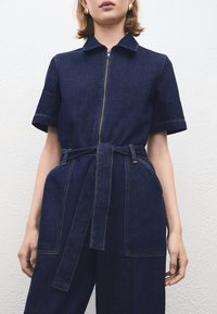 Tmavomodrý rifľový overal s krátkymi rukávmi, predným zipsom a opaskom v páse. Má dve veľké predné vrecká a výrazné švy.