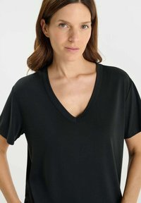 Vrouw met lichte huid en bruin haar, gekleed in een eenvoudige zwarte V-hals t-shirt met korte mouwen, staande tegen een effen lichte achtergrond.