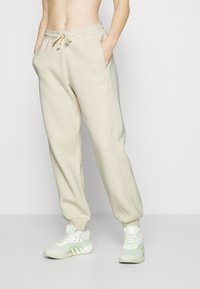 Pantalones de chándal beige con cintura ajustable, bolsillos laterales y puños acanalados. Los pantalones presentan un logo sutil en un lado, combinados con zapatillas deportivas verdes.