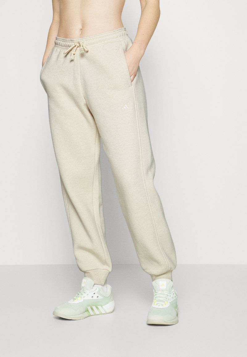 Pantalones de chándal beige con cintura ajustable, bolsillos laterales y puños acanalados. Los pantalones presentan un logo sutil en un lado, combinados con zapatillas deportivas verdes.