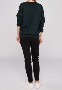 Osoba stojící zády k nám, oblečená v tmavém dlouhém rukávu, černých kalhotách a bílých teniskách, na jednobarevném světlém pozadí.