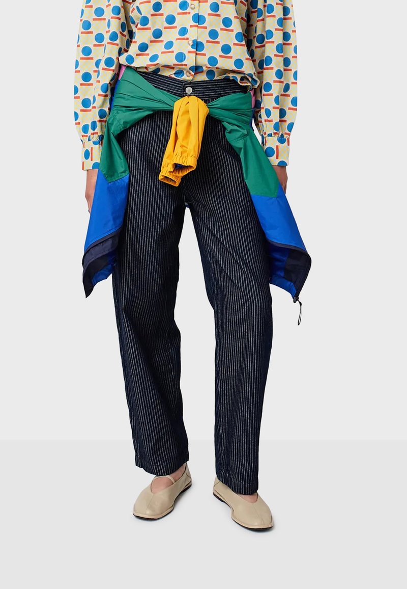 Persona che indossa pantaloni neri a righe sottili, una camicia a fantasia geometrica multicolore, ballerine beige e una giacca verde, blu e gialla annodata in vita.