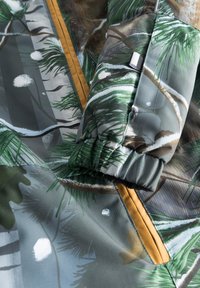 Kravejakke med et grønt og gråt grantræsmønster med hvide sneakkenter; inkluderer en guldlås og elastiske manchetter.