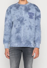 Persona che indossa una felpa Tommy Jeans tie-dye blu con la scritta "Tommy 1985 Jeans" e jeans grigi, in piedi contro uno sfondo chiaro.