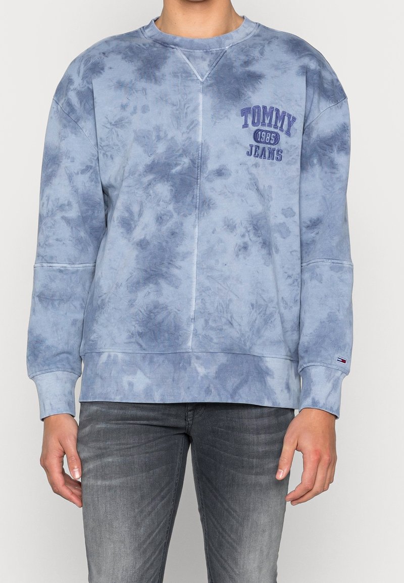 Persona che indossa una felpa Tommy Jeans tie-dye blu con la scritta "Tommy 1985 Jeans" e jeans grigi, in piedi contro uno sfondo chiaro.