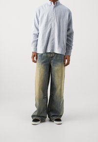 Hombre vestido con una camisa de botones azul claro y pantalones vaqueros anchos de color azul desvanecido y amarillo, con zapatillas negras y blancas, de pie sobre un fondo liso.