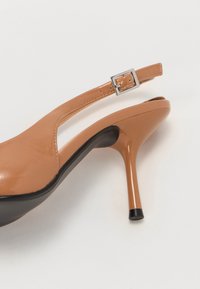 Escarpin ouvert en cuir verni beige avec un haut talon noir, doté d'une bride de cheville et d'un détail de boucle carrée en argent.