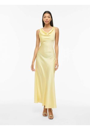 Kvinne står og har på seg en gulblek, ermeløs satengkjole som går ned til gulvet, med fallende halslinning og gullfargede åpne tåhæler på hvit bakgrunn.