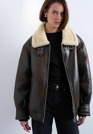 Joven mujer vestida con jeans negros y una chaqueta de piel marrón oscuro con un gran cuello color crema, de pie contra un fondo liso.