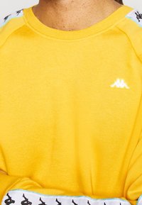 Felpa gialla con logo bianco sul petto e loghi neri sulle strisce bianche delle spalle e dei polsini, indossata da una persona con le braccia incrociate.