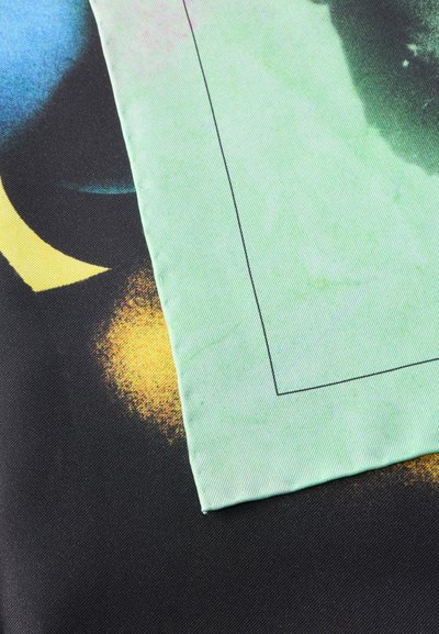Κοντινή λήψη διπλωμένου ανοιχτό πράσινου υφάσματος με γαζωμένη περιγραφή, τοποθετημένο πάνω σε μαύρο ύφασμα με κίτρινα και μπλε αφηρημένα μοτίβα.