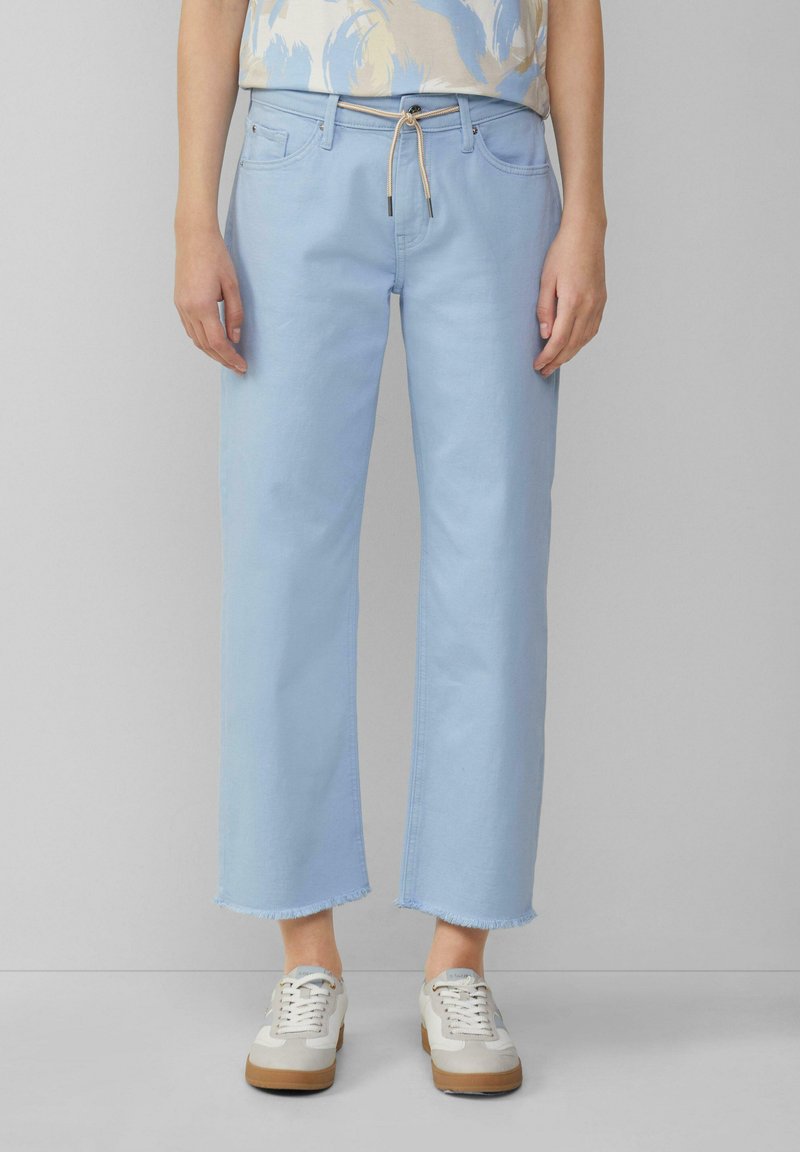 Jeans anchos de mezclilla azul claro con dobladillo deshilachado y cintura con cordón, combinados con zapatillas blancas y beige.
