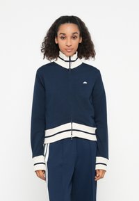 Marineblå zip-up hettegenser med høy krage og kremstripete mansjetter, med en liten logo på brystet og en tettsittende midje.