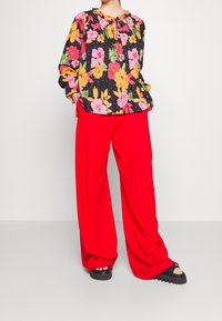 La blusa floreale presenta fiori rosa intenso, arancioni e verdi su uno sfondo nero; abbinata a pantaloni larghi rossi a gamba larga e sandali neri con zeppa.