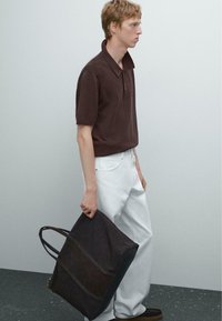 Polo shirt marrone con colletto a zip, pantaloni bianchi e borsa a tracolla in pelle scura. La borsa ha due manici e una texture liscia.