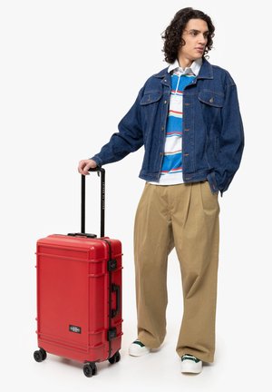 Maleta rígida roja con acentos y mango negros, cuatro ruedas y un candado. El usuario lleva puesta una chaqueta de mezclilla, una camiseta a rayas y pantalones beiges.