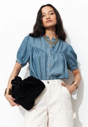 Femme aux longs cheveux foncés, portant un chemisier bleu à manches courtes, un pantalon blanc taille haute, des bijoux dorés, tenant une grande pochette noire à volants.