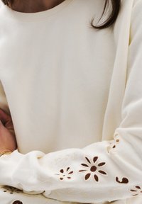 Maglione bianco con maniche lunghe, caratterizzato da ricami floreali ritagliati nei polsini. Il tessuto sembra morbido con una texture liscia.