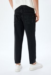 Vaqueros de denim negro con un corte relajado, con dos bolsillos traseros y costuras sutiles. Combinados con zapatillas blancas sobre un fondo neutro.