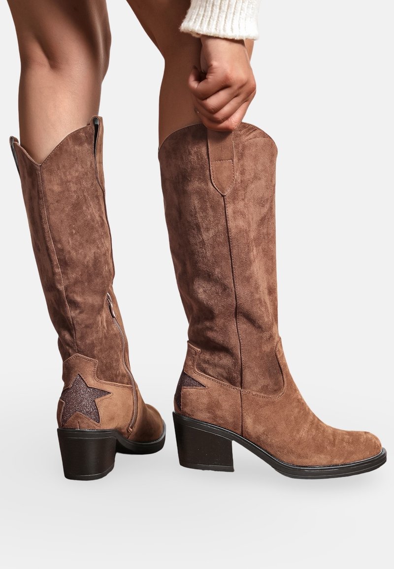 Bottes hautes en daim marron avec un léger bout pointu, fermeture éclair sur le côté et une étoile noire en accent sur le talon. La semelle à bloc assure une stabilité.