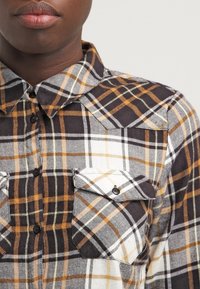 Camisa de cuadros en tonos negro, blanco y naranja. Cuenta con cierre frontal de botones, cuello y dos bolsillos en el pecho con solapas. Tela texturizada.