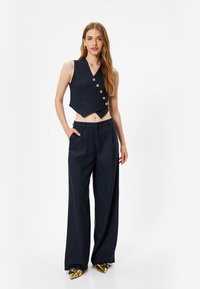 Marineblaues, gestreiftes, kurzes Vest mit goldenen Knöpfen, kombiniert mit weiten Hosen. Der Stoff hat eine glatte Textur und eine taillierte Passform. Goldene, spitz zulaufende Schuhe.